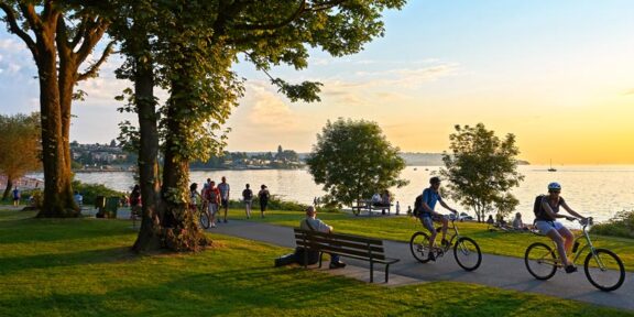 City park with people biking along a path and the ocean behind them at sunset.